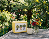 A yellow vintage-style hard case with a white front panel and illustrated figures holding flower bouquets sits on a wooden table outdoors. A ceramic mug and a small glass vase of mixed flowers are beside it, with a lush garden background of green foliage and yellow flowers.
